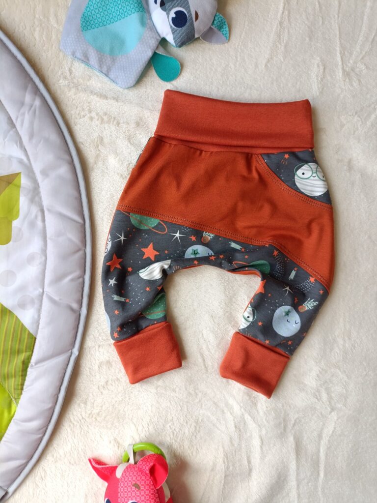 Sarouel évolutif pour bébé, avec empiècement orange brique et jambes en tissu gris anthracite à motif spatial, composé de planètes, fusées et étoiles. Large ceinture et revers orange brique, repliés pour illustrer sa taille minimale.