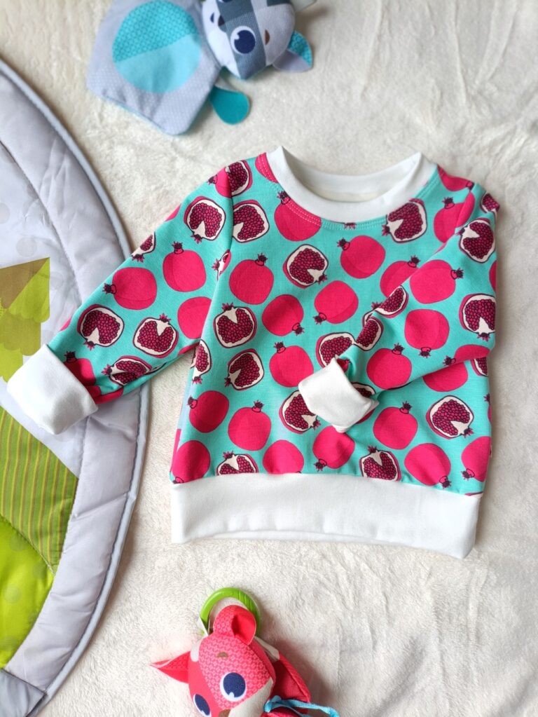 Tee-shirt évolutif pour enfant à manches longues, en tissu écru à motif de grenades roses sur un fond turquoise, avec encolure, poignets et bande de taille noirs. Présenté suspendu sur un cintre, avec les poignets repliés pour illustrer sa taille minimale.