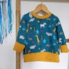 Tee-shirt évolutif pour enfant à manches longues, en tissu bleu canard à motif d'animaux de la savane, avec encolure, poignets et bande de taille moutarde. Présenté suspendu sur un cintre, avec les poignets repliés pour illustrer sa taille minimale.
