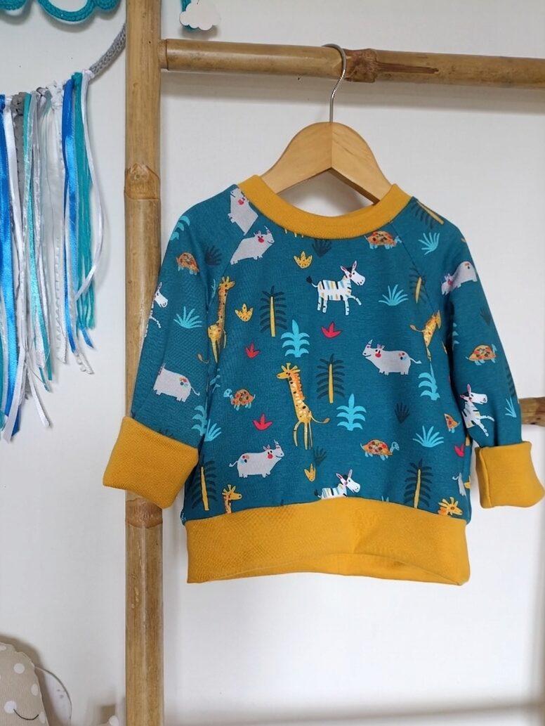Tee-shirt évolutif pour enfant à manches longues, en tissu bleu canard à motif d'animaux de la savane, avec encolure, poignets et bande de taille moutarde. Présenté suspendu sur un cintre, avec les poignets repliés pour illustrer sa taille minimale.