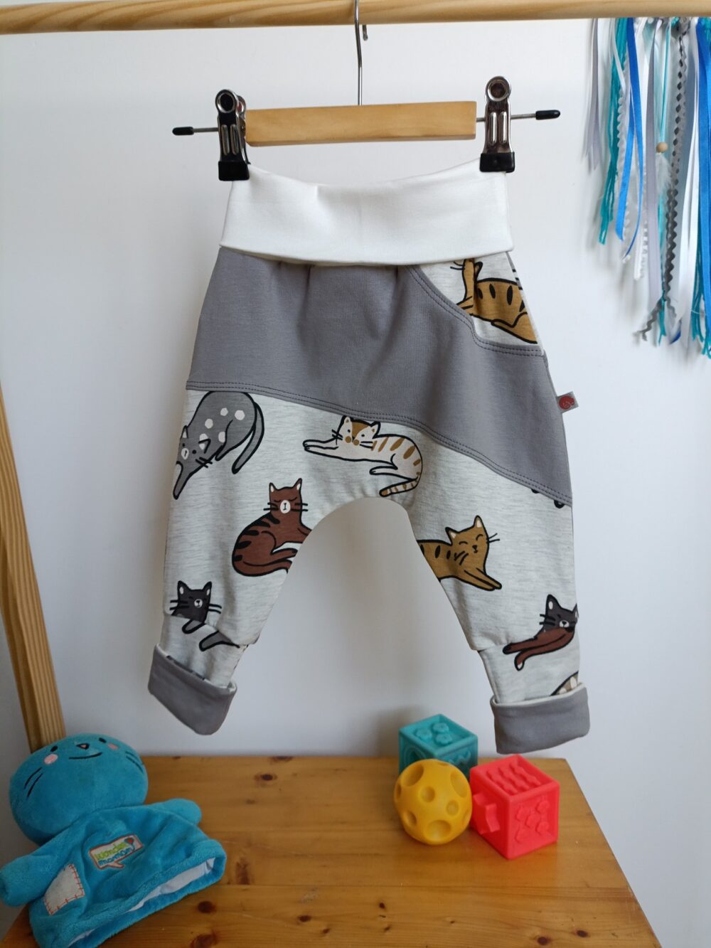 Sarouel évolutif pour bébé à motif de chats sur fond gris chiné, avec empiècement gris, large ceinture blanche et revers gris. Présenté avec la ceinture et les revers repliés pour illustrer sa taille minimale.