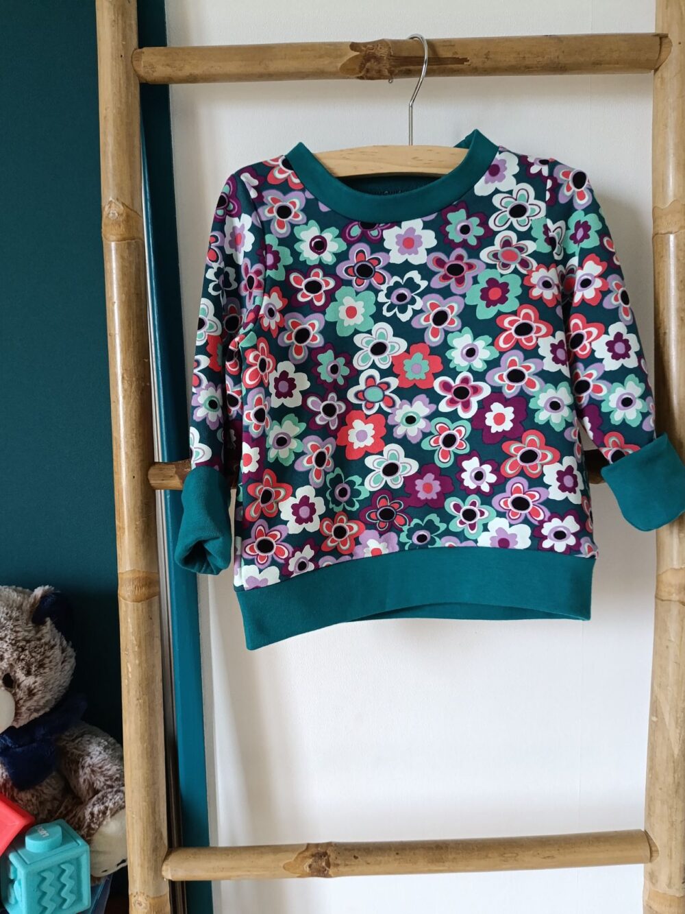Sweat évolutif pour bébé à manches longues, en tissu à motif de fleurs multicolores sur fond bleu pétrole, avec encolure, poignets et bande de taille bleu pétrole. Présenté suspendu sur un cintre.