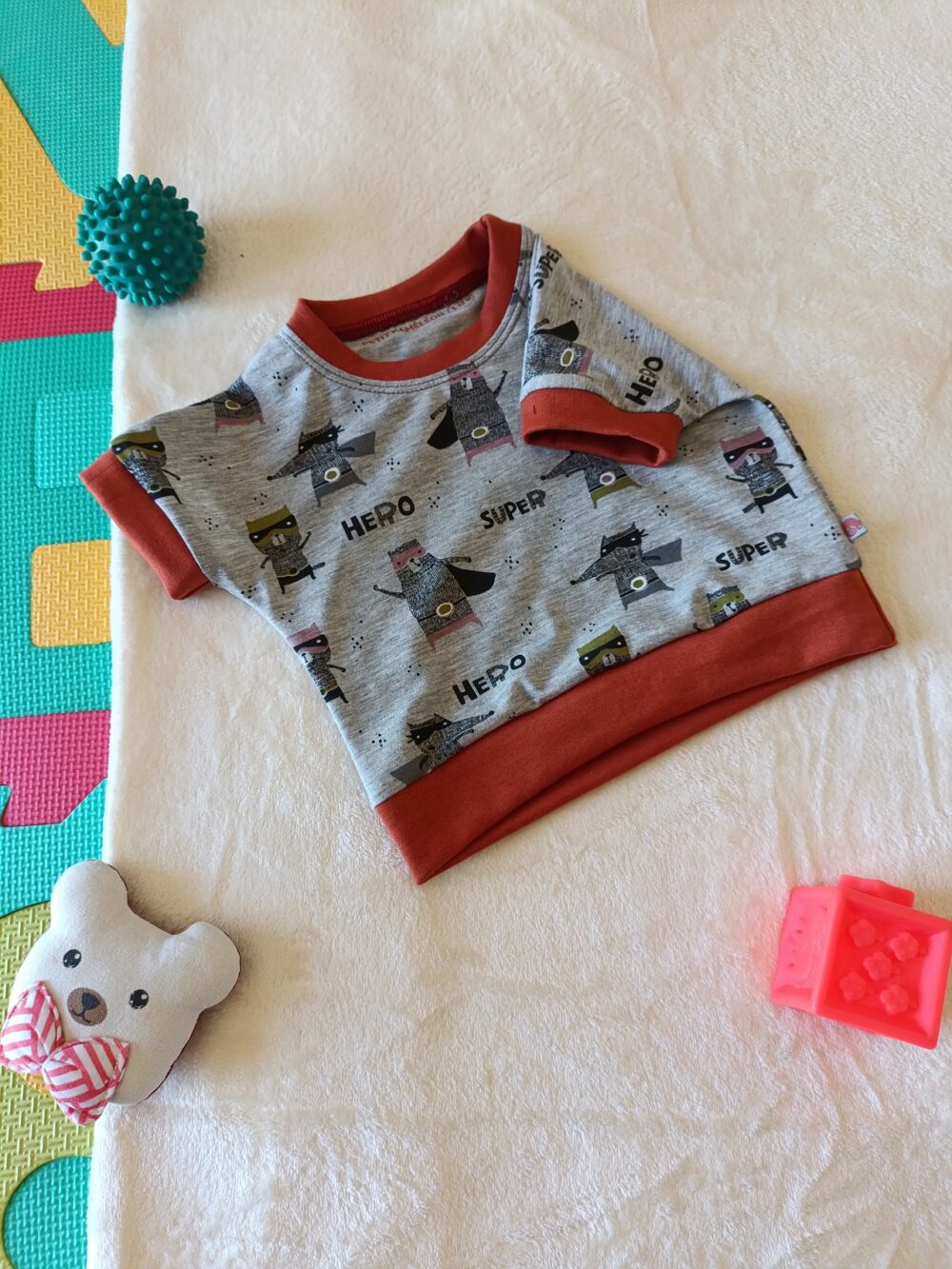 Tee-shirt évolutif pour enfant à manches courtes, en tissu gris chiné à motif de super-héros, avec encolure, poignets et bande de taille orange brique.