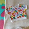 Tee-shirt évolutif pour bébé à manches courtes, en tissu blanc à motif abstrait multicolore. Présenté entièrement déplié pour montrer sa taille maximale.