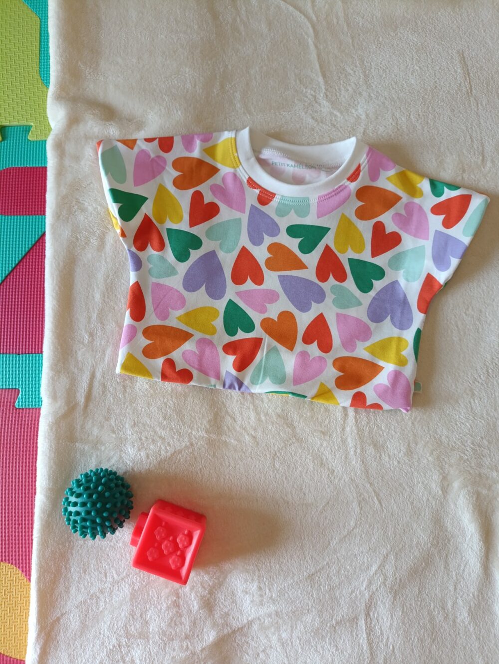 Tee-shirt évolutif pour bébé à manches courtes, en tissu blanc à motif de cœurs multicolores. Présenté avec les bandes repliées, correspondant à sa taille la plus petite.