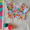 Tee-shirt évolutif pour bébé à manches courtes, en tissu blanc à motif de cœurs multicolores. Présenté avec les bandes repliées, correspondant à sa taille la plus petite.