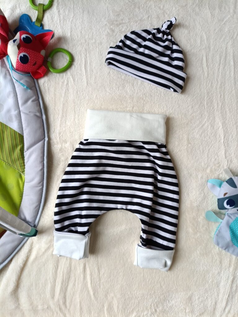 Ensemble évolutif pour bébé composé d'un bonnet et d'un pantalon à rayures horizontales noires et blanches. Le bonnet est doté d'un nœud sur le dessus et d'un large revers blanc. Le pantalon présente une large ceinture et des revers blancs, repliés pour illustrer sa taille minimale.