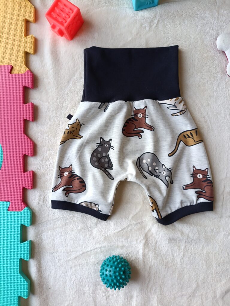 Short évolutif pour bébé à motif de chats gris, roux et beiges sur fond écru, avec large ceinture et revers bleu marine. Présenté avec la ceinture dépliée pour illustrer sa taille maximale.