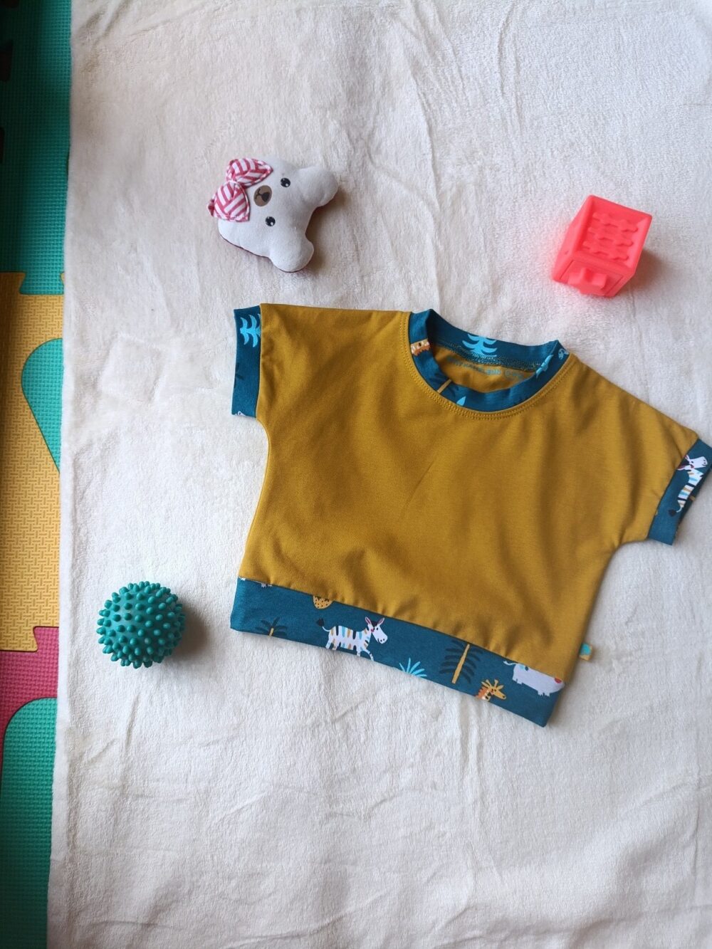 Tee-shirt pour bébé couleur moutarde avec encolure et bas de manches en tissu bleu à motifs. Le modèle présente une poche poitrine contrastante et une bande décorative bleue à motifs sur le bas du vêtement.
