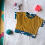 Tee-shirt pour bébé couleur moutarde avec encolure et bas de manches en tissu bleu à motifs. Le modèle présente une poche poitrine contrastante et une bande décorative bleue à motifs sur le bas du vêtement.