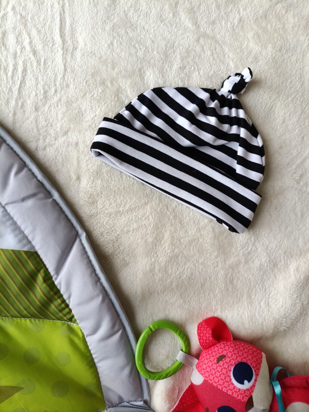 Striped black-and-white baby hat with a knot on top, lying on a cream rug beside a green play mat and a pink plush toy nearby.