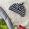 Striped black-and-white baby hat with a knot on top, lying on a cream rug beside a green play mat and a pink plush toy nearby.