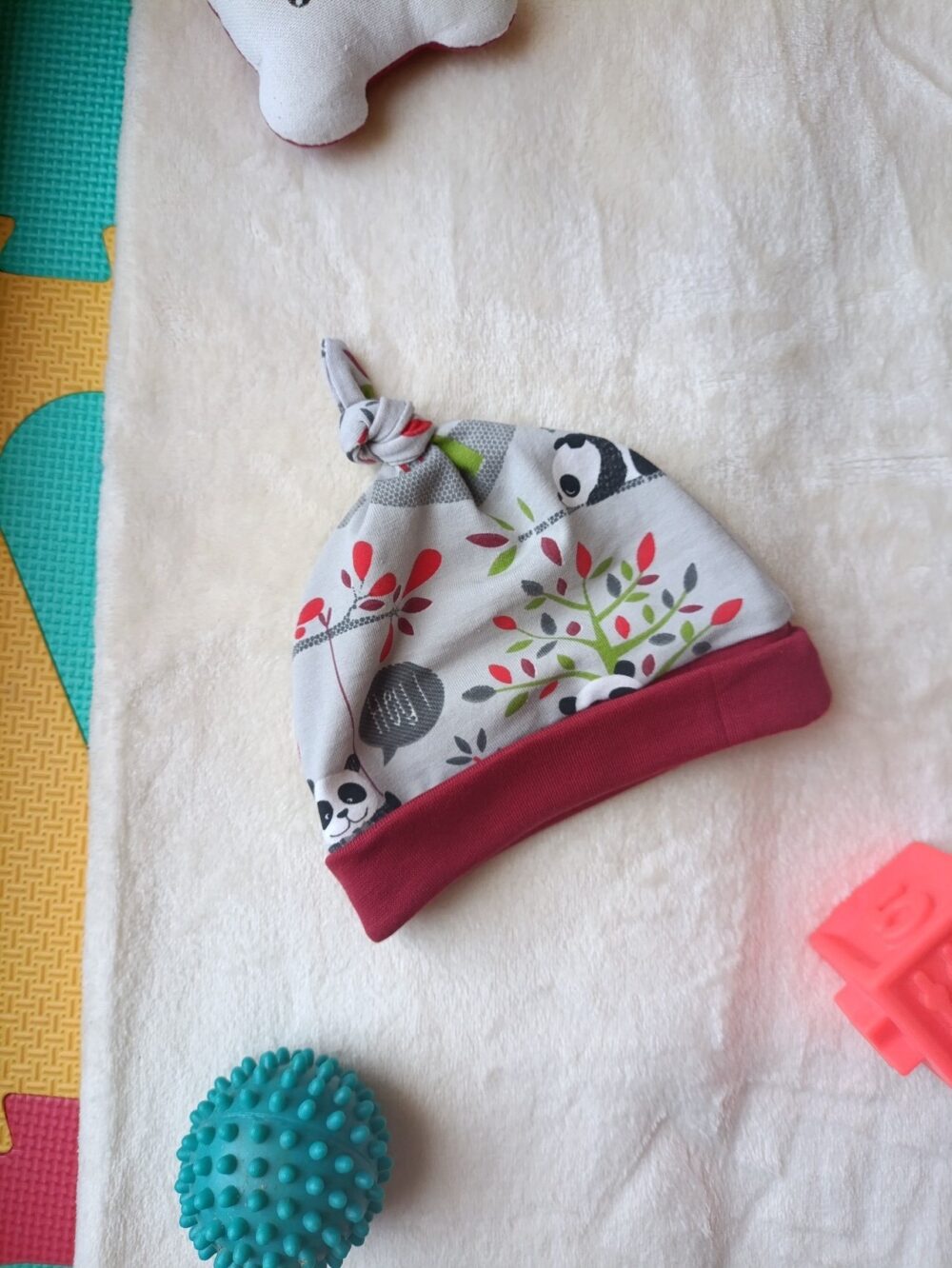 Baby hat with panda print and red brim resting on a soft white towel, with a teal spiky ball nearby