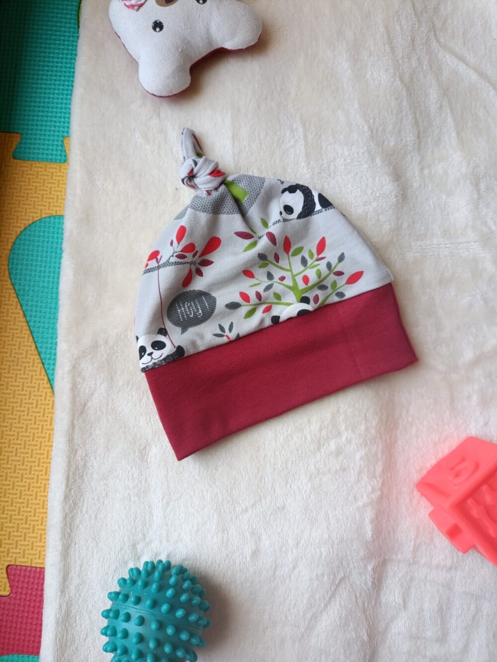 Knitted baby hat with panda print and red band on a soft light surface, with teal textured ball and pink blocks nearby.