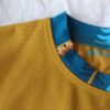Close-up of a mustard-yellow fabric with teal collar trim and visible stitching, showing part of a patterned tag reading 'PETIT K'.