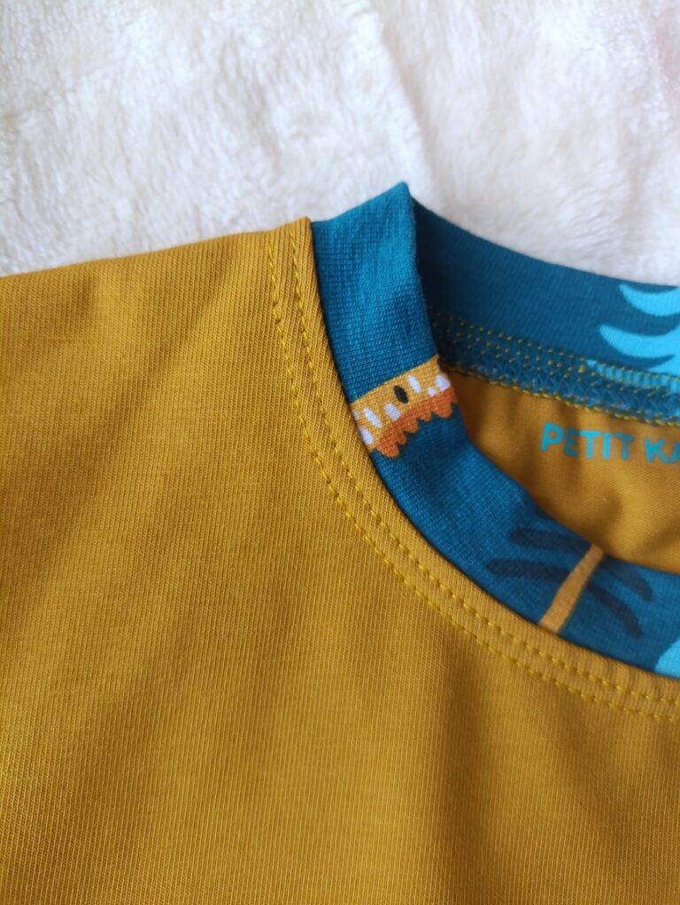 Gros plan sur l'encolure d'un tee-shirt moutarde, avec bande d'encolure en tissu bleu à motif animaux savane assorti.