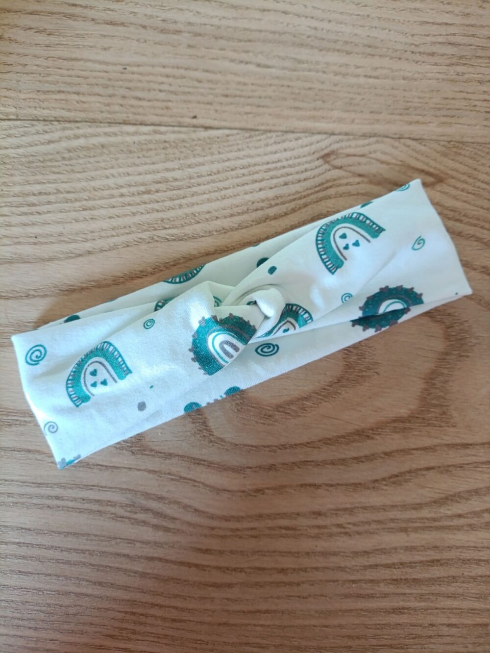 Bandeau en tissu blanc à motif cachemire vert canard, avec un nœud plat au centre. Modèle large de style croisé, présenté à plat sur un fond en bois clair.