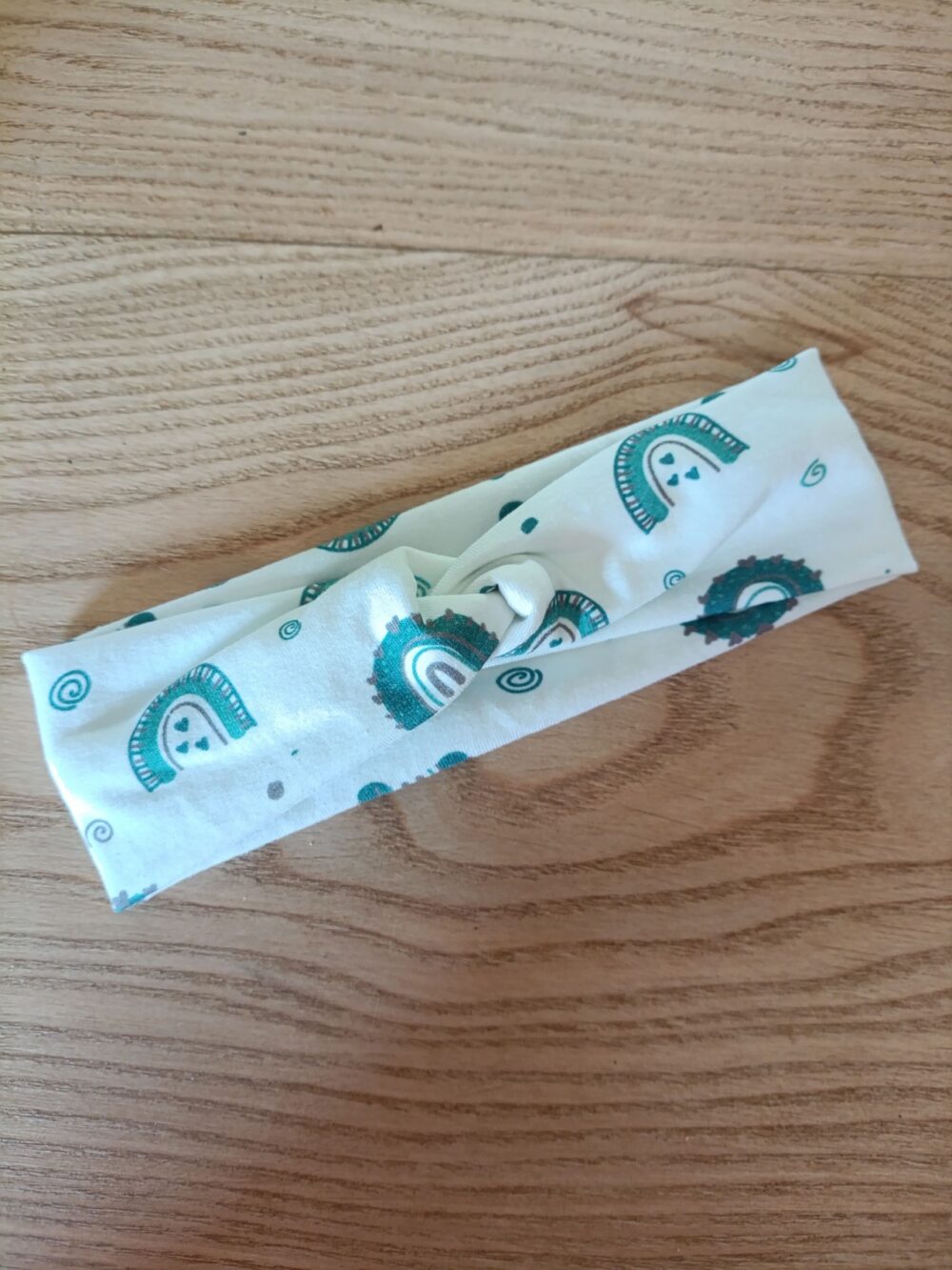 Bandeau en tissu blanc à motif cachemire vert canard, avec un nœud plat au centre. Modèle large de style croisé, présenté à plat sur un fond en bois clair.