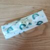 Bandeau en tissu blanc à motif cachemire vert canard, avec un nœud plat au centre. Modèle large de style croisé, présenté à plat sur un fond en bois clair.