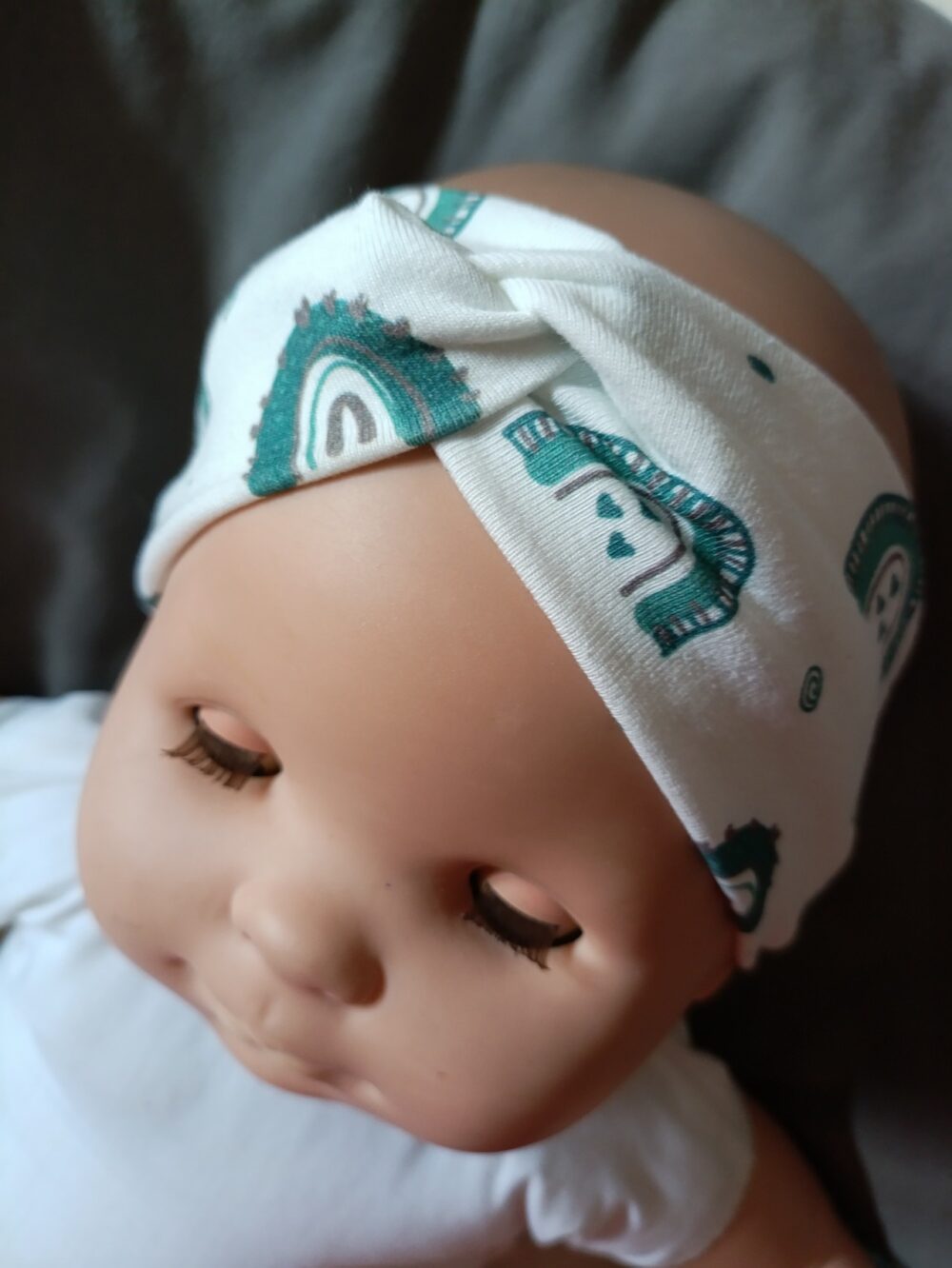 Bandeau large en tissu blanc à motif arcs en ciel verts, croisé sur le devant. Présenté porté sur une poupée.