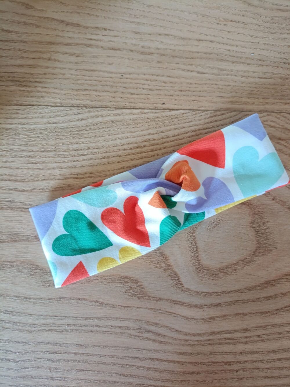 Bandeau en tissu blanc à motif coeurs, avec un nœud plat au centre. Modèle large de style croisé, présenté à plat sur un fond en bois clair.