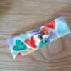 Bandeau en tissu blanc à motif coeurs, avec un nœud plat au centre. Modèle large de style croisé, présenté à plat sur un fond en bois clair.
