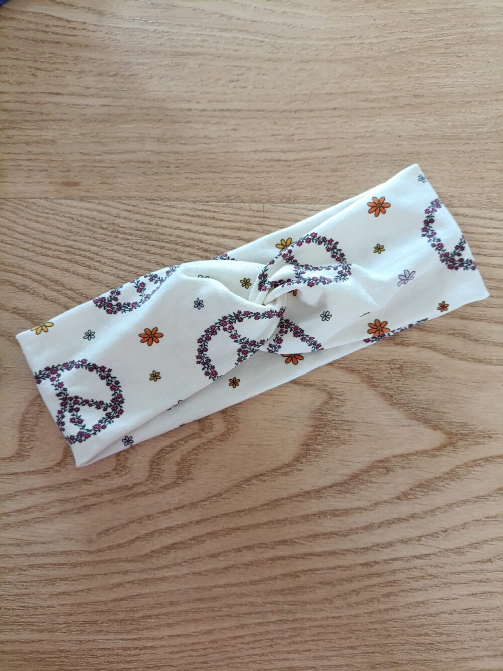Bandeau en tissu blanc à motifs Peace & love, avec un nœud plat au centre. Modèle large de style croisé, présenté à plat sur un fond en bois clair.
