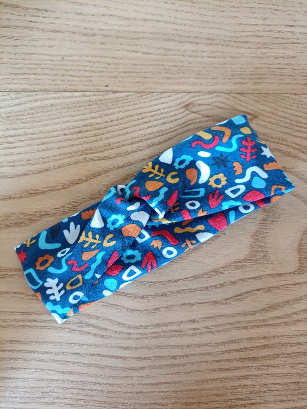 Bandeau en tissu coton à motifs abstraits multicolores, avec un nœud plat au centre. Modèle large de style croisé, présenté à plat sur un fond en bois clair.