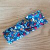 Bandeau en tissu coton à motifs abstraits multicolores, avec un nœud plat au centre. Modèle large de style croisé, présenté à plat sur un fond en bois clair.