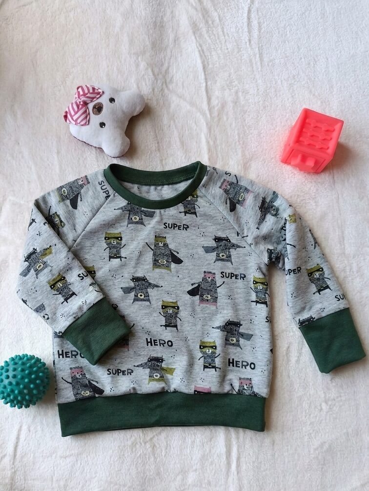 Tee-shirt évolutif pour enfant à manches longues, en tissu gris chiné à motif de super-héros, avec encolure, poignets et bande de taille vert sapin. Présenté avec les poignets dépliés pour illustrer sa taille maximale.