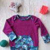 Tee-shirt évolutif pour enfant à manches longues, violet, avec encolure américaine et poche kangourou et finitions en tissu à motif végétal bleu, turquoise et violet. Présenté avec les poignets dépliés pour illustrer sa longueur maximale.