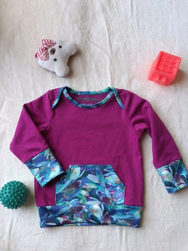 Tee-shirt évolutif pour enfant à manches longues, violet, avec encolure américaine et poche kangourou et finitions en tissu à motif végétal bleu, turquoise et violet. Présenté avec les poignets dépliés pour illustrer sa longueur maximale.