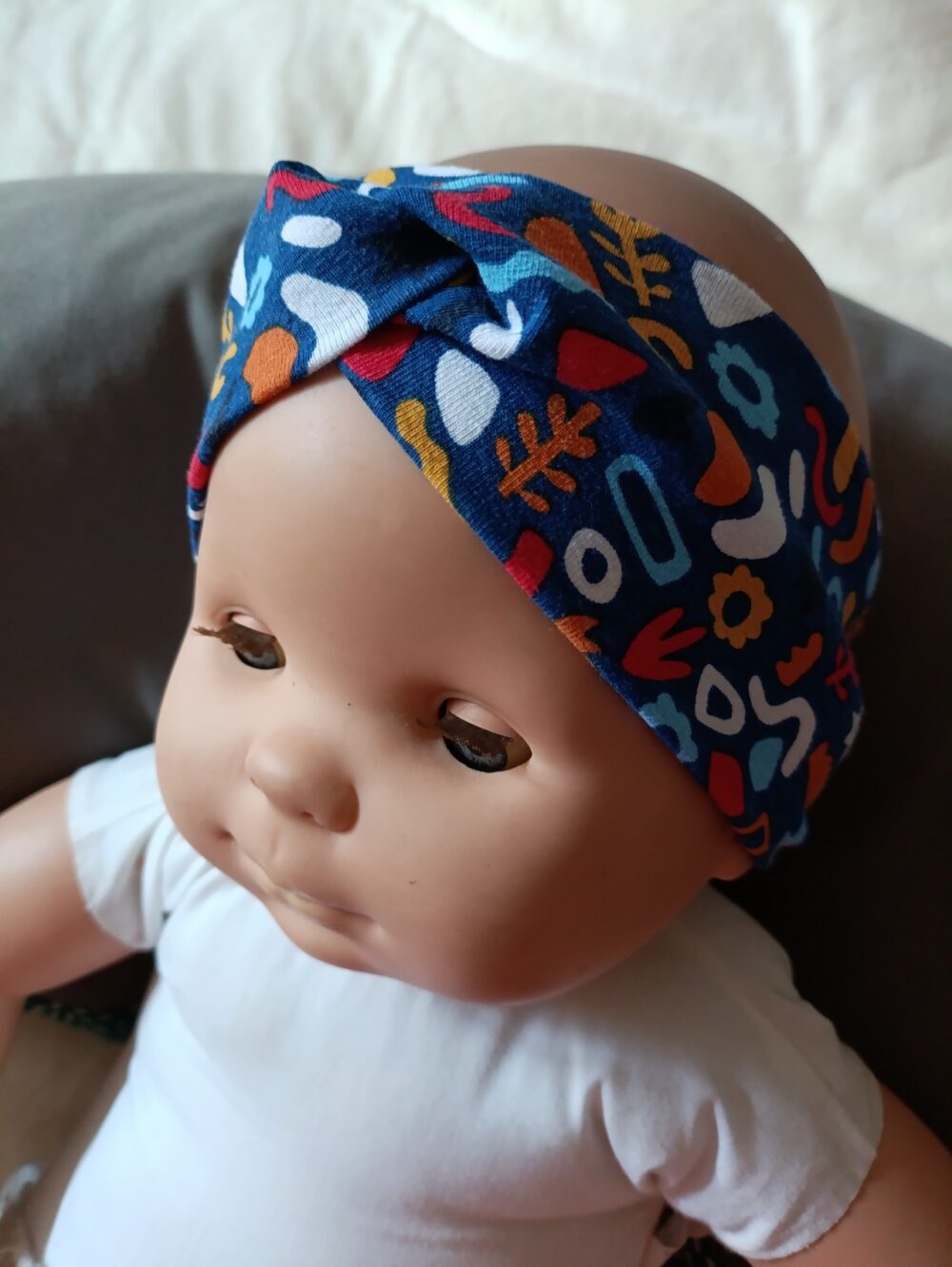 Bandeau large en tissu bleu marine à motif abstrait multicolore, croisé sur le devant. Le motif mêle des formes géométriques et végétales en bleu clair, blanc, rouge, orange et ocre. Présenté porté sur une poupée.