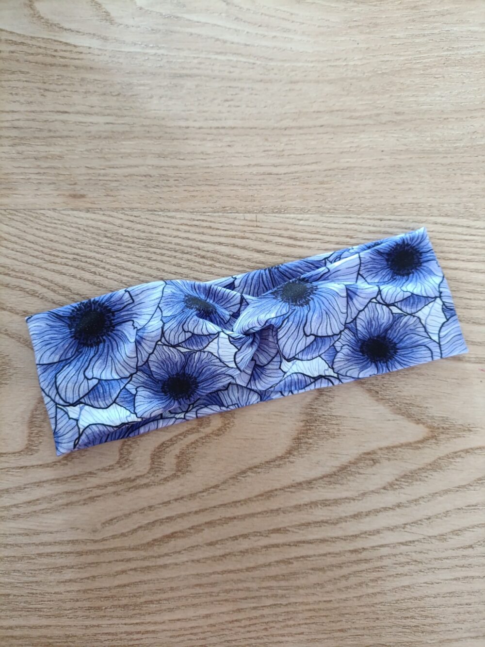 Bandeau large en tissu blanc à motif floral bleu et violet, représentant de grandes fleurs aux pétales fins. Modèle à bande large, présenté à plat sur un fond en bois clair.