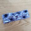Bandeau large en tissu blanc à motif floral bleu et violet, représentant de grandes fleurs aux pétales fins. Modèle à bande large, présenté à plat sur un fond en bois clair.